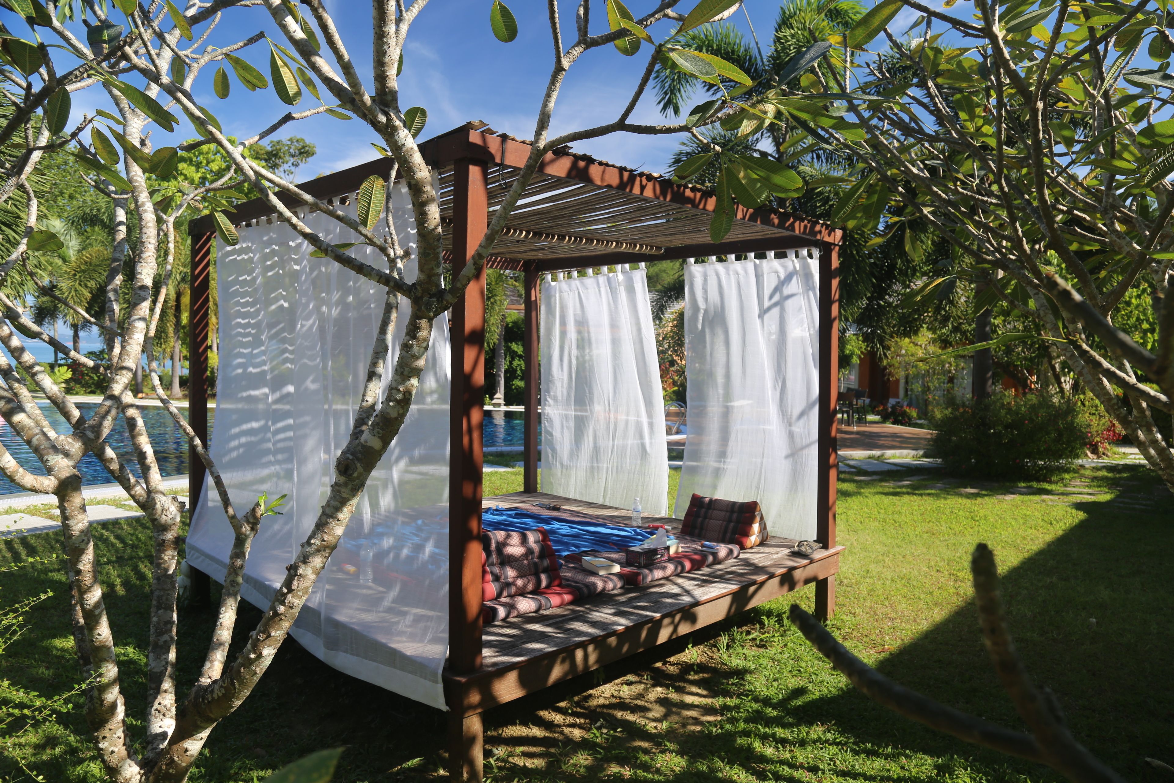 A wooden outdoor seating area with a canopy, surrounded by trees and a pool in the background.