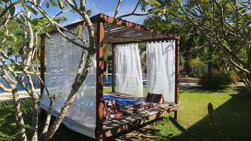 A wooden outdoor seating area with a canopy, surrounded by trees and a pool in the background.