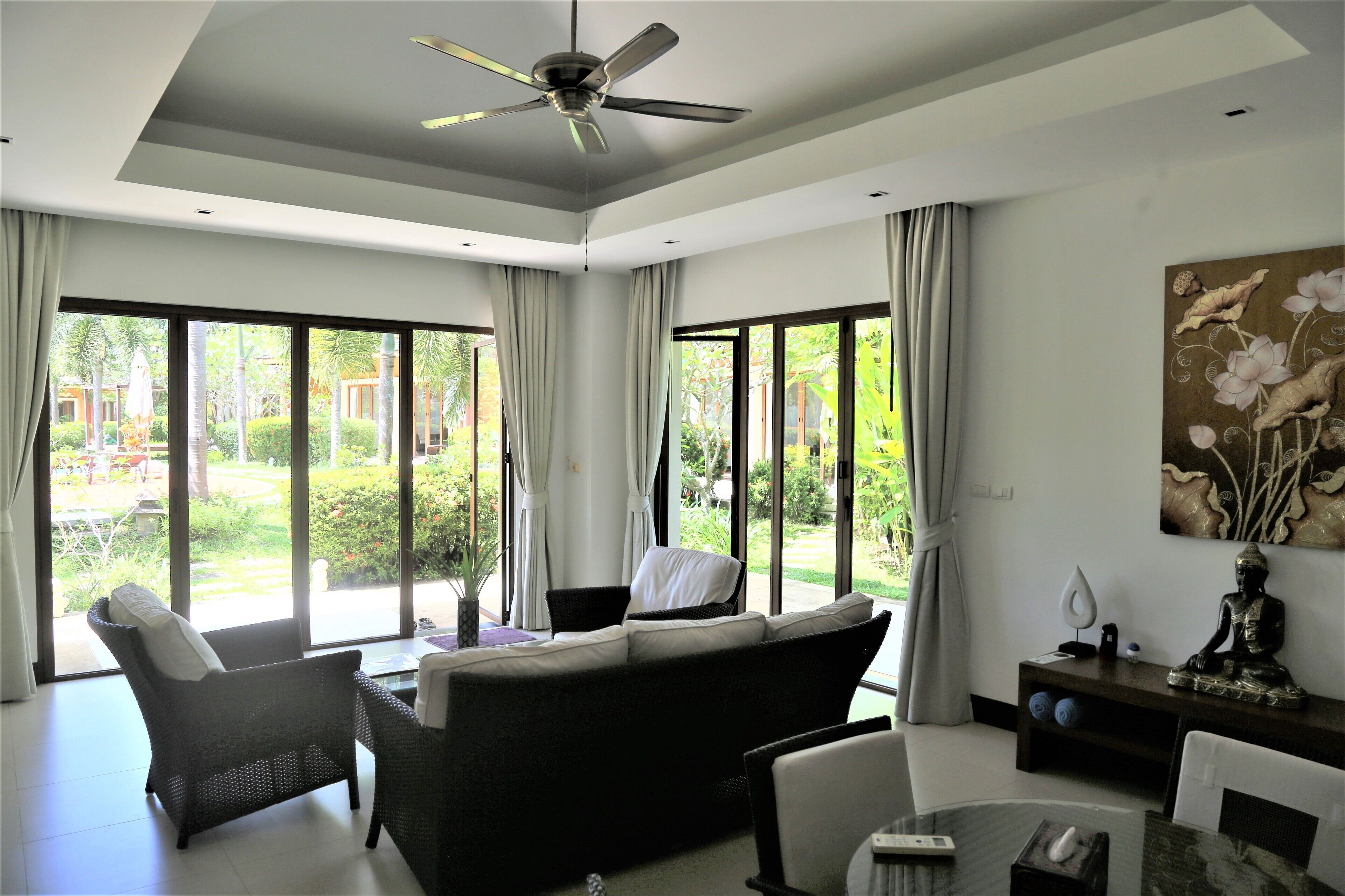 A modern living room with a sofa, chairs, a ceiling fan, large windows, and a Buddha statue.