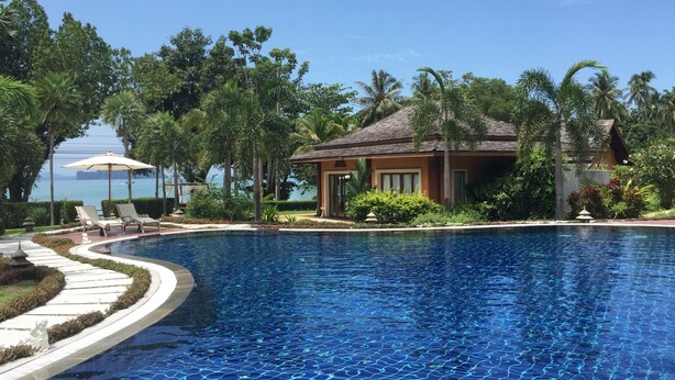 A swimming pool with a tiled deck, surrounded by palm trees and a building with a tiled roof.