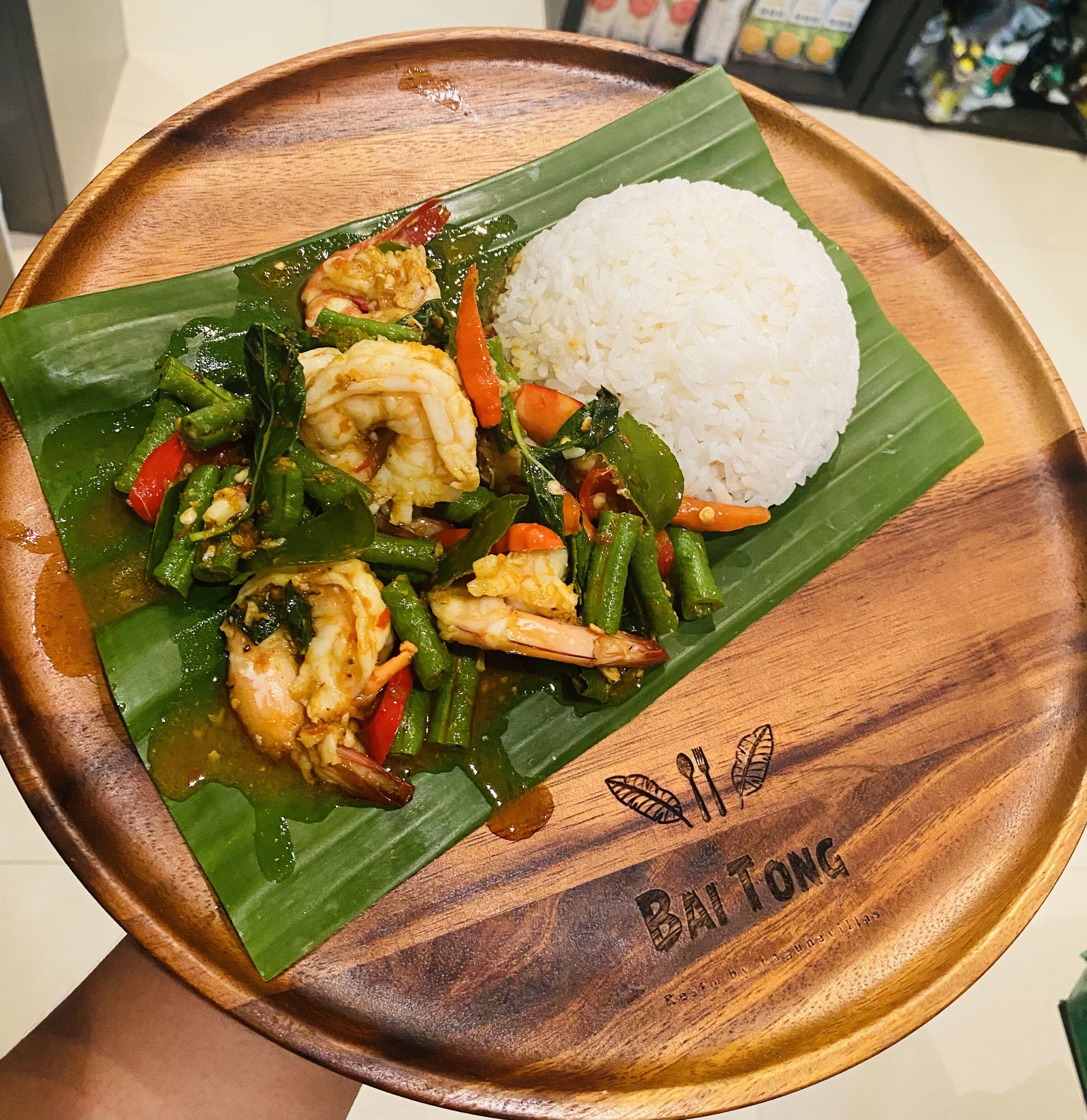 Un plato de madera con hoja de plátano, que contiene un guiso de camarones y verduras, junto con una porción de arroz blanco.