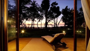 A room with a view of a sunset over palm trees, a lounge chair, and a glass wall.