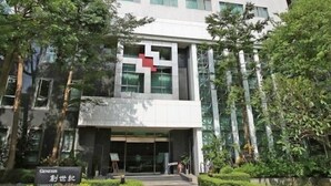 Un edificio moderno de varios pisos con una señal roja prominente de cruz, rodeado de follaje bien cuidado y un cielo despejado.