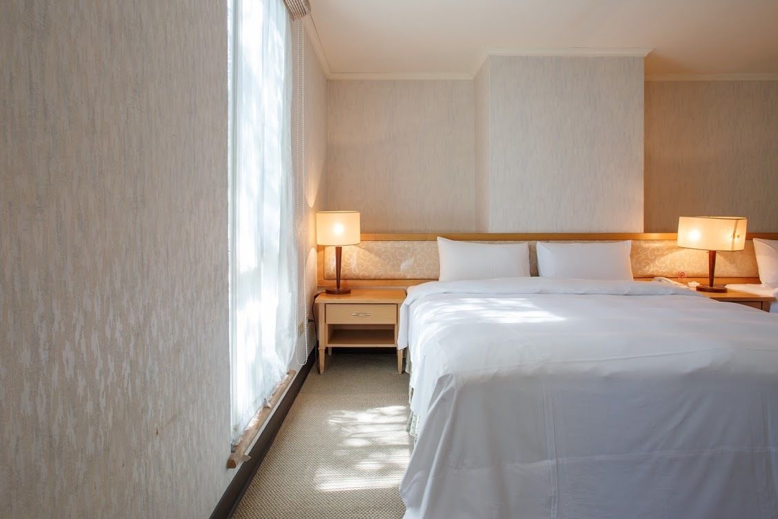 Quarto de hotel com uma cama grande, duas mesas de cabeceira com luminárias, um criado-mudo e uma janela com cortinas translúcidas.