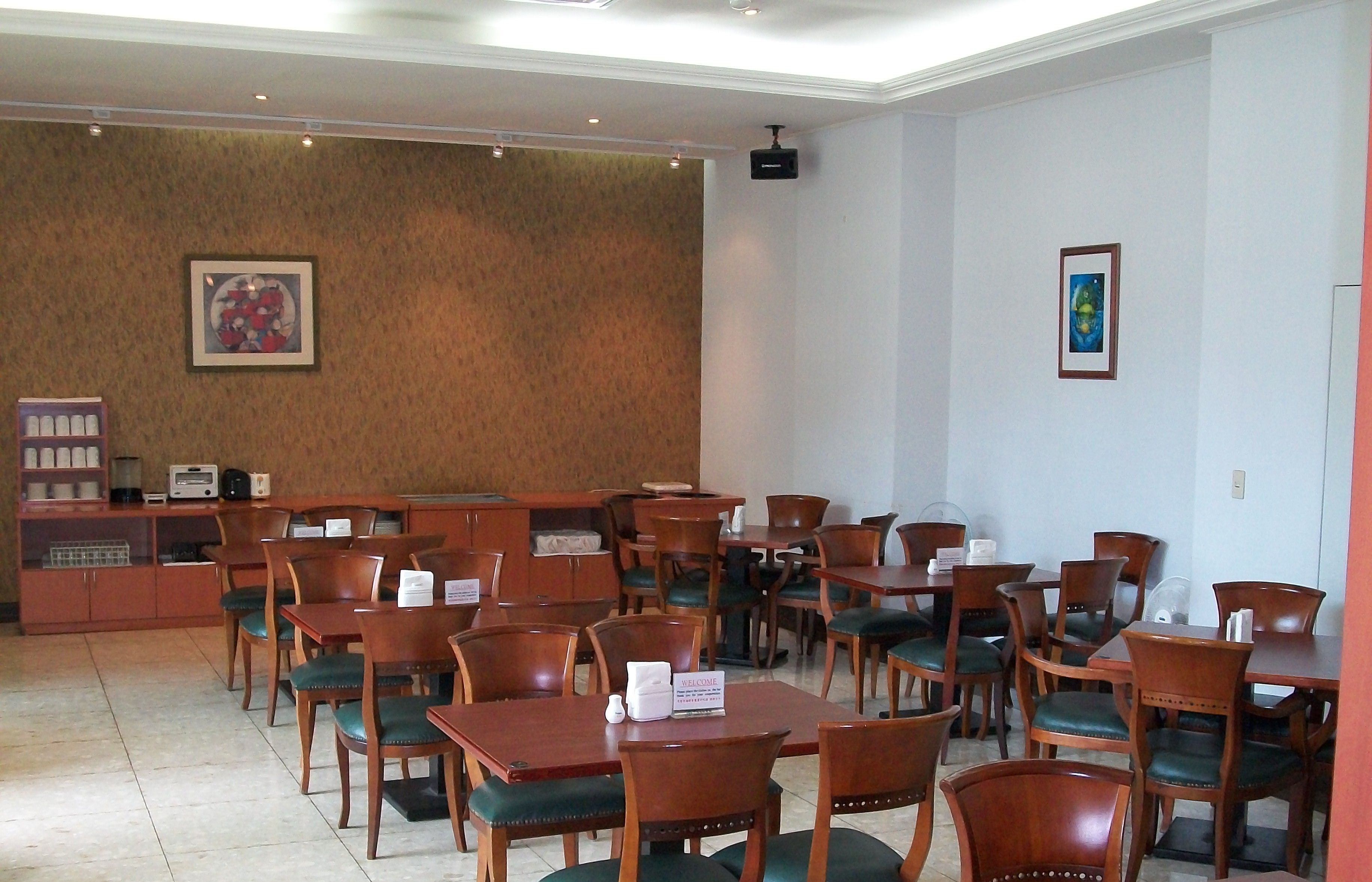 A hotel lobby with wooden tables and chairs, a reception desk, and a wall-mounted television.