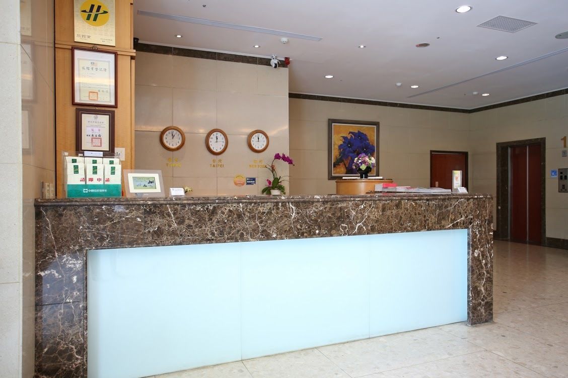 Un mostrador de recepción de hotel con encimera de mármol, relojes, diplomas enmarcados y un arreglo floral.