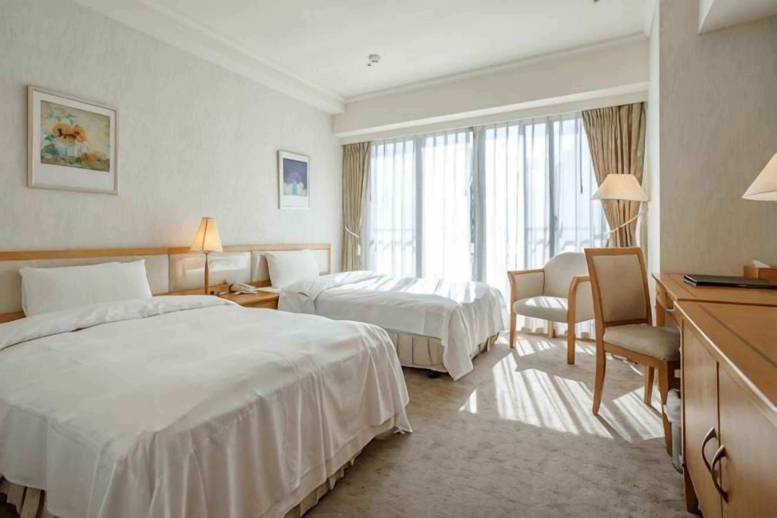 Une chambre d’hôtel avec deux lits, un bureau, une chaise, une lampe et une grande fenêtre avec des rideaux.