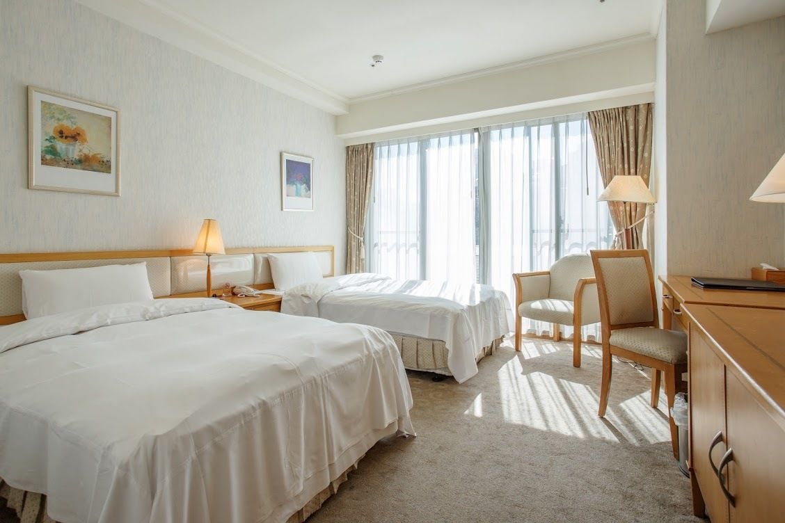 Ein Hotelzimmer mit zwei Betten, einem Schreibtisch, einem Stuhl und einem fensterbelaubten Fenster.