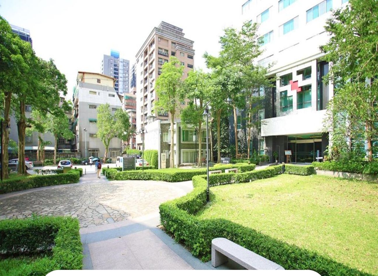 A well-maintained hotel courtyard with neatly trimmed hedges, a stone pathway, and modern buildings in the background.