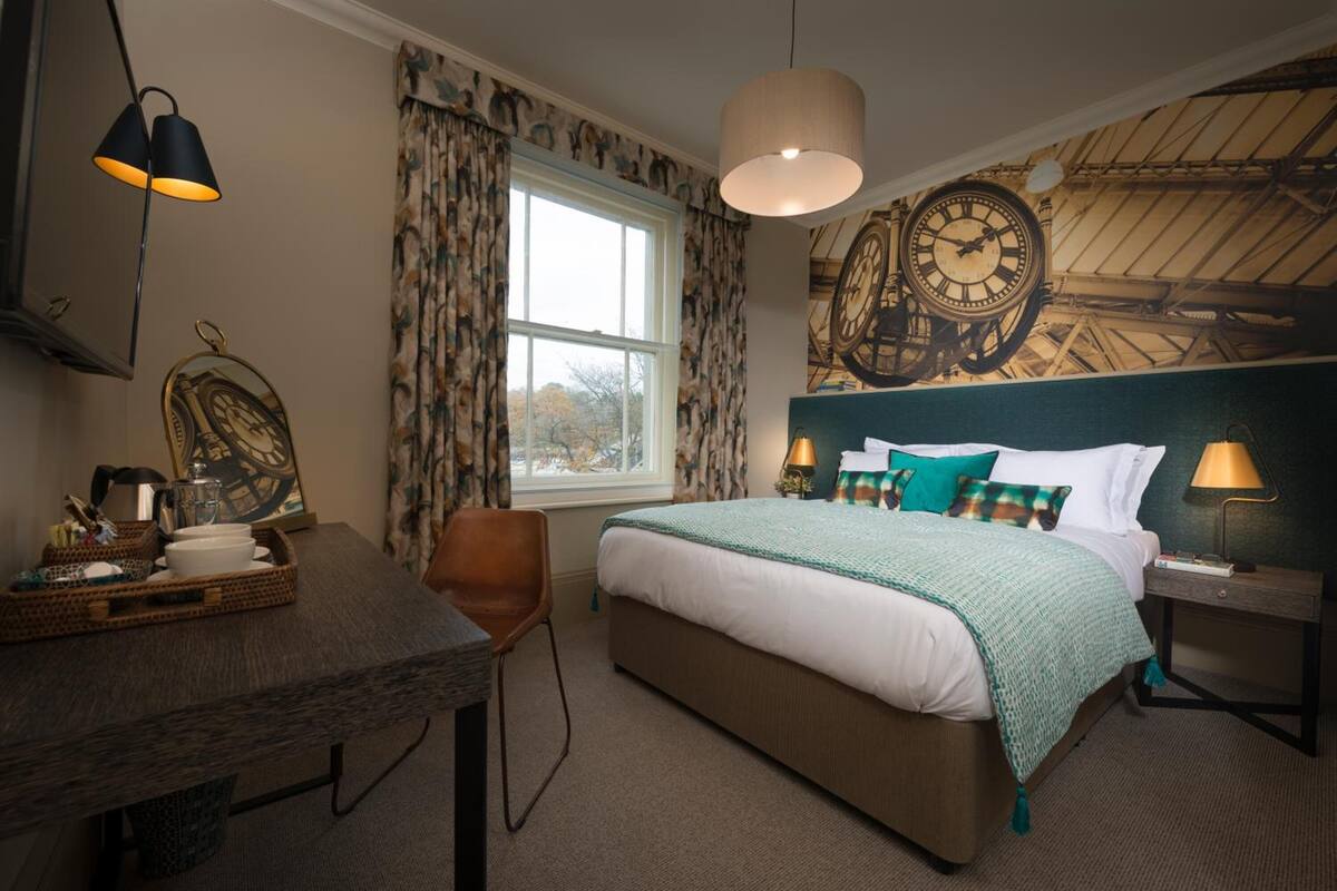 A hotel room with a large bed, a desk with a lamp, a chair, and a window with curtains.
