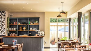 L’intérieur d’un restaurant est aménagé avec des tables et des chaises en bois, un coin bar doté d’étagères et de grandes fenêtres qui laissent entrer la lumière naturelle.