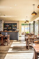 Interior de restaurante con mesas y sillas de madera, una zona de bar con estanterías y grandes ventanales que dejan entrar luz natural.