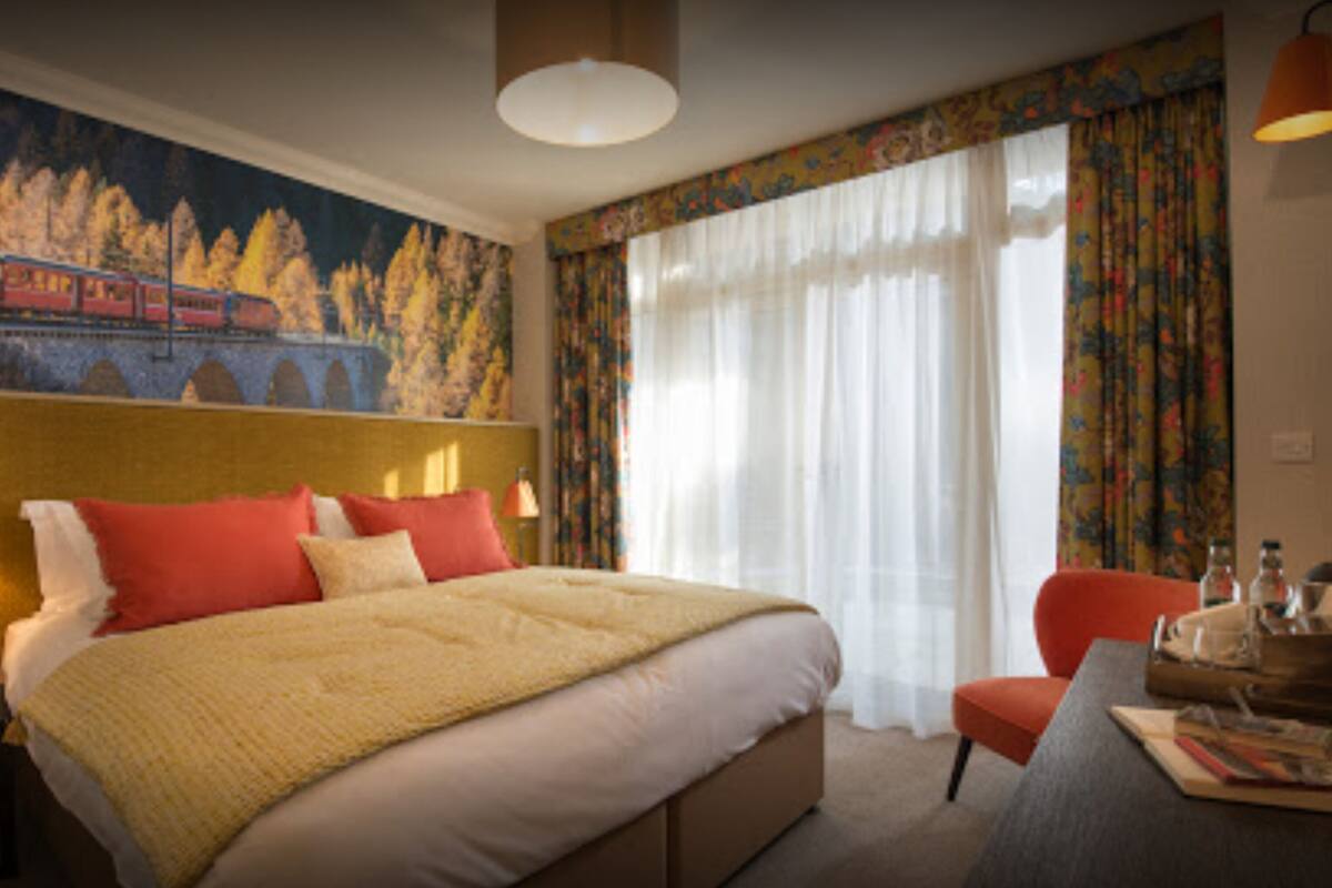 A hotel room with a large bed, a desk, a chair, a lamp, and a painting of a train on the wall.