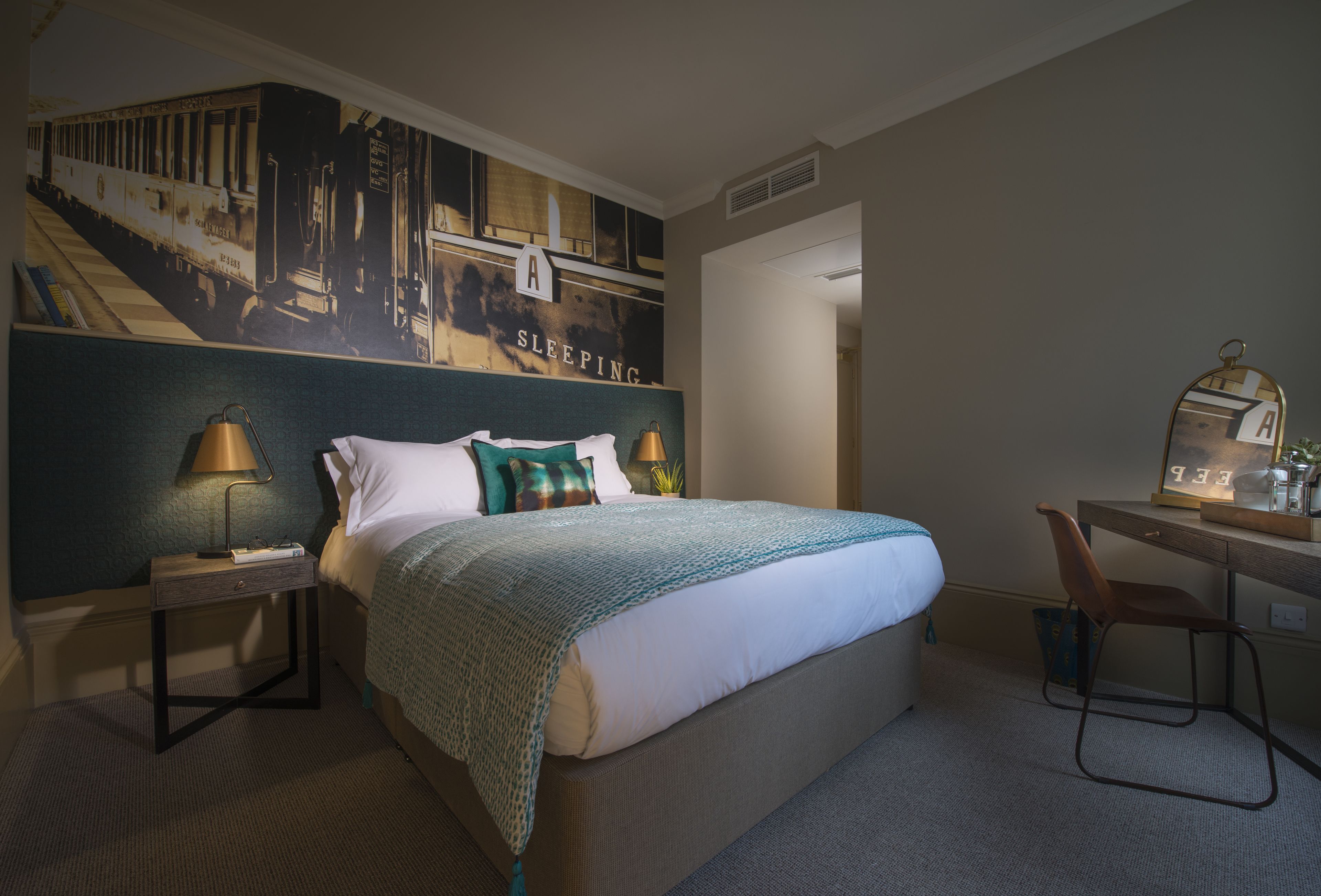 A hotel room with a large bed, a desk with a chair, a nightstand with a lamp, and a wall mural.