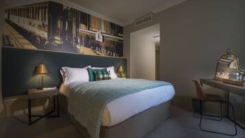 A hotel room with a large bed, a desk with a chair, a nightstand with a lamp, and a wall mural.