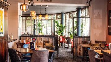 Un restaurante con varias mesas y sillas, una zona de bar y grandes ventanales con plantas.