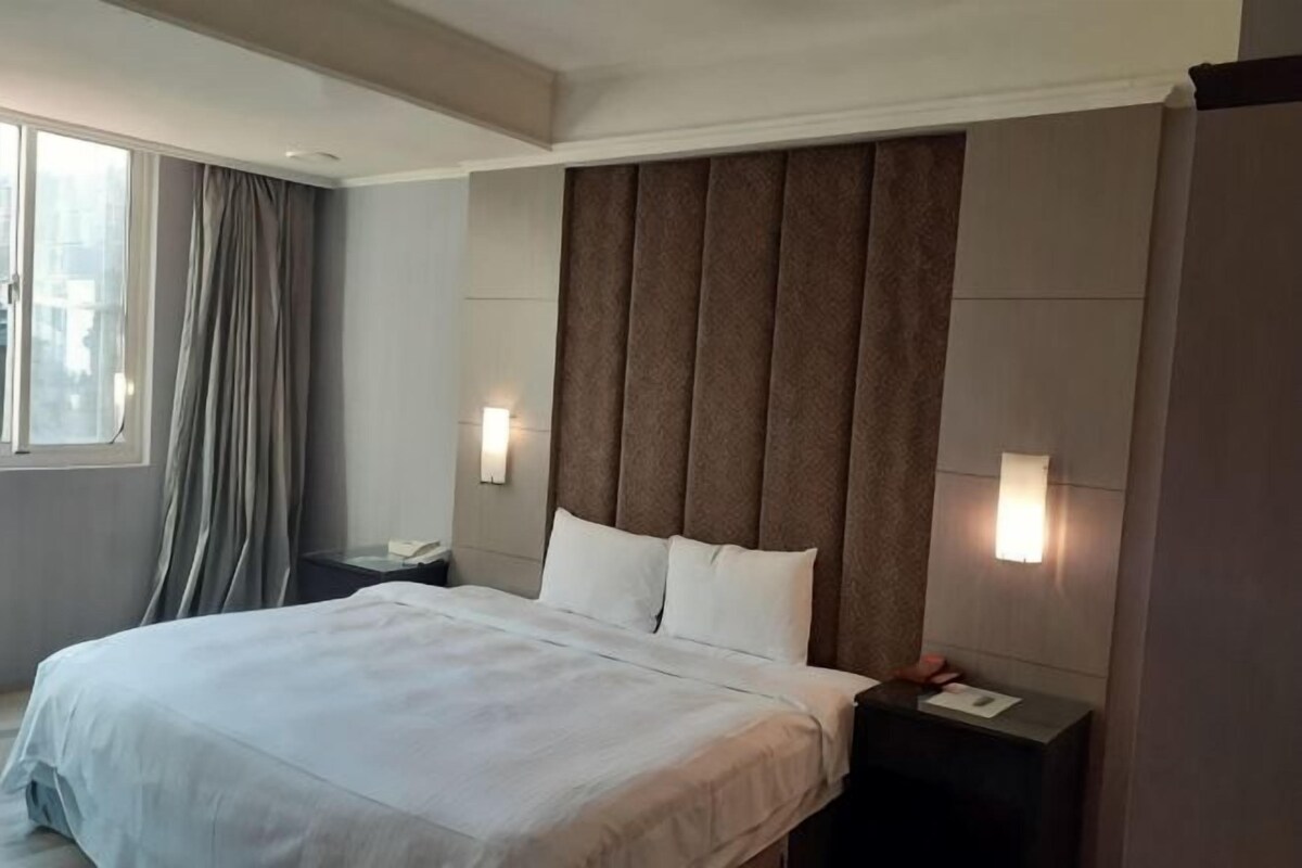 Une chambre d’hôtel comprenant un lit, une table de chevet, une lampe, une fenêtre avec des rideaux et un luminaire fixé au mur.