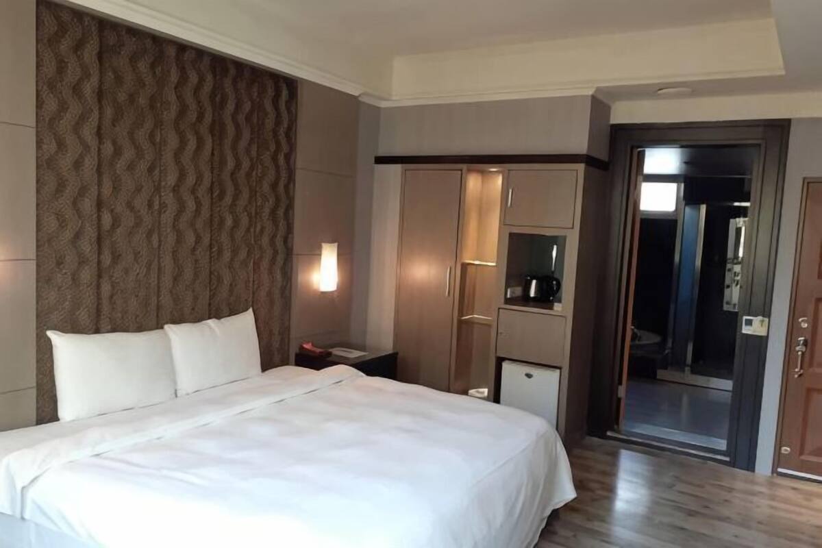 A hotel room with a large bed, a wardrobe, a desk with a lamp, and a sliding door leading to a bathroom.