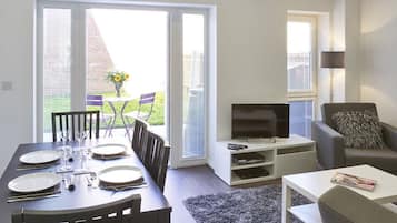 Un comedor con una mesa dispuesta para cuatro personas, un sofá, un televisor y una mesita con una revista.