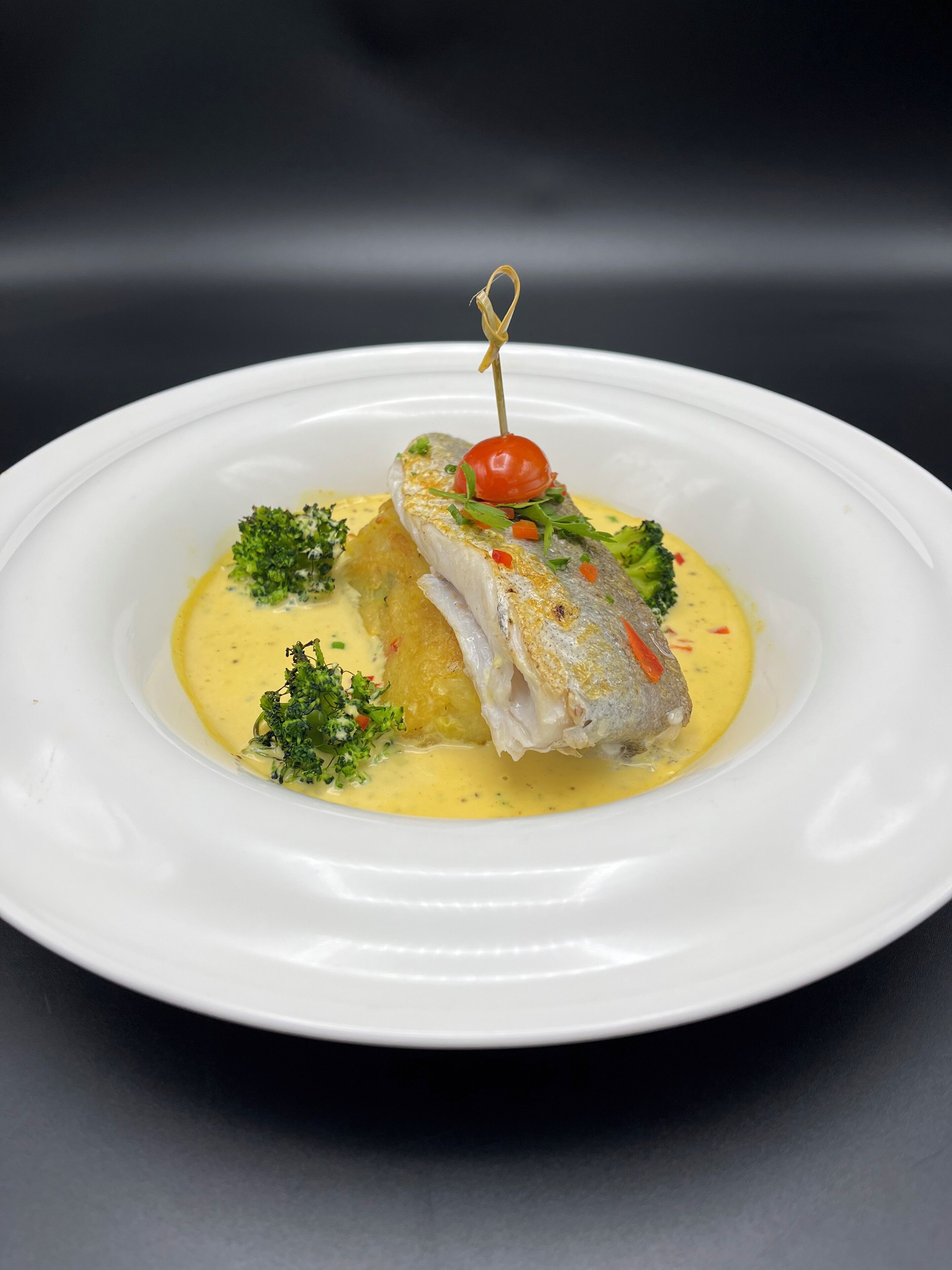 A white plate with a fish fillet, a cherry tomato, and broccoli on a dark background.