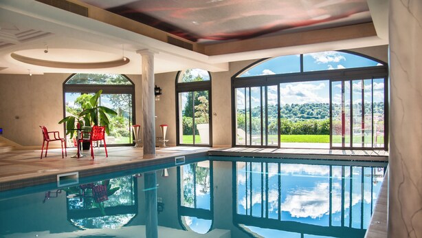 An indoor swimming pool with clear views of the outdoors, including a garden and distant hills.