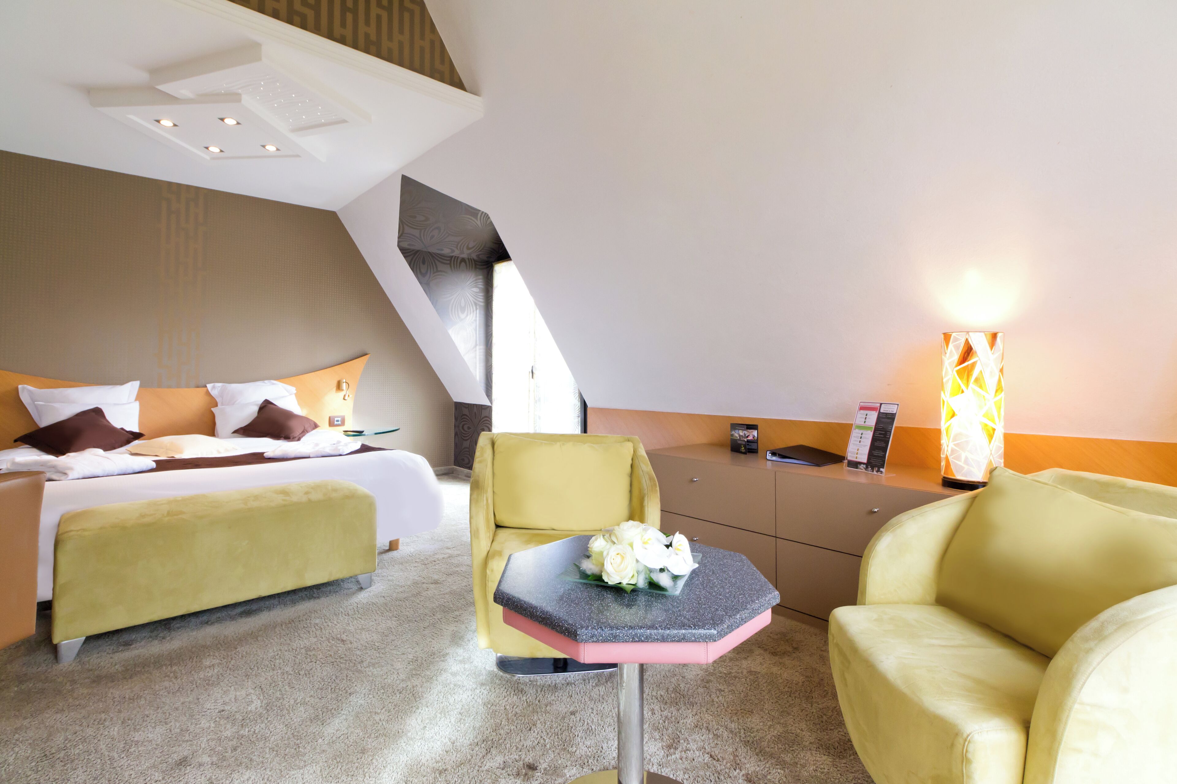 A hotel room with a bed, a yellow armchair, a small table with a bouquet of flowers, and a bedside table with a lamp.