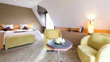 A hotel room with a bed, a yellow armchair, a small table with a bouquet of flowers, and a bedside table with a lamp.