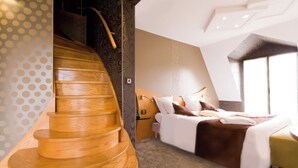 A modern hotel room with a wooden spiral staircase, a bed with white and brown bedding, and a large window providing natural light.