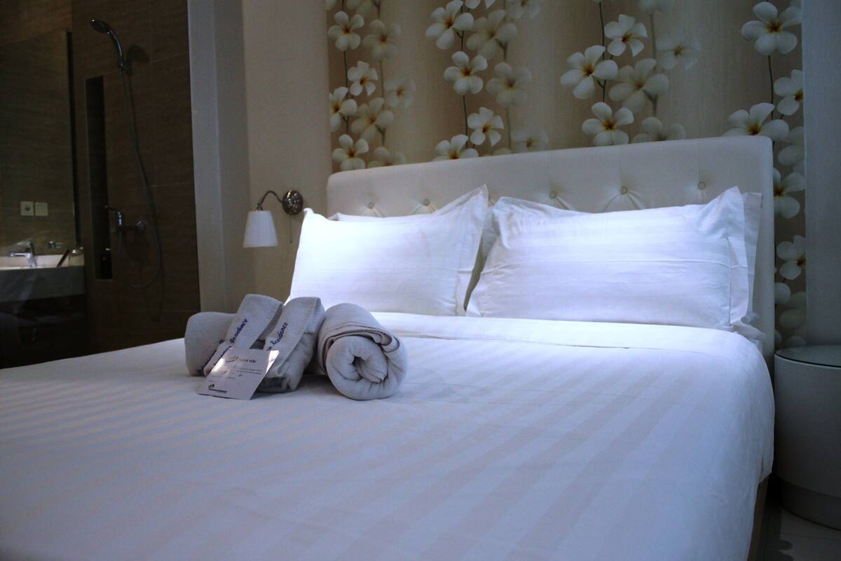 A neatly made bed with white linens and pillows, a folded towel, and a towel rack.
