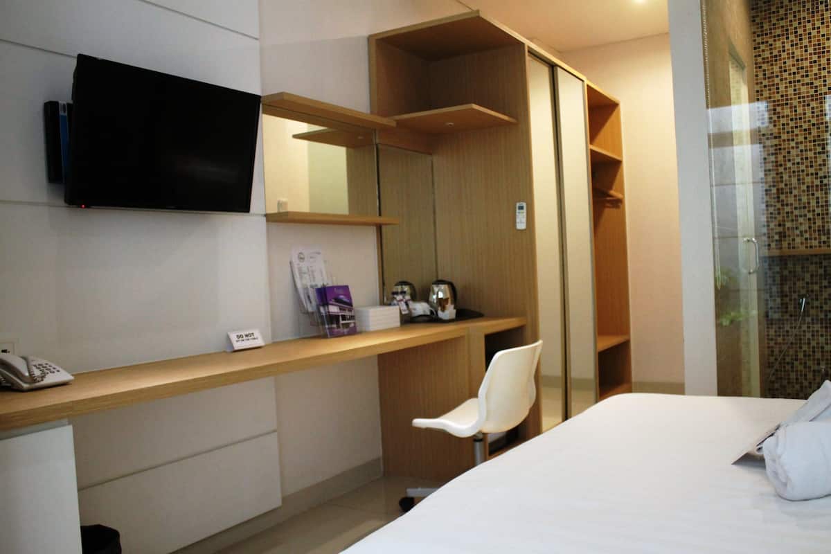 A hotel room with a bed, a desk with a telephone, a chair, a television mounted on the wall, and a shower area with a glass door.