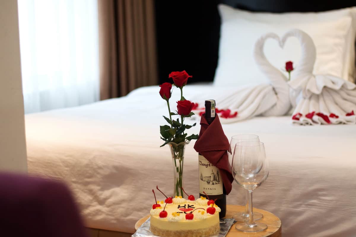 Un lit avec du linge de lit blanc, un coussin en forme de cœur, un bouquet de roses rouges, une bouteille de vin, un verre et un gâteau aux cerises.