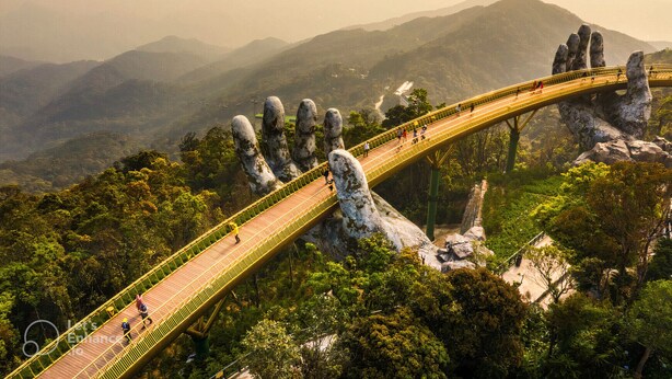 Un puente con un dise ño singular, sostenido por grandes formaciones rocosas, rodeado de exuberante vegetación y montañas al fondo.