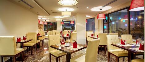 A restaurant with marble flooring, beige chairs, and red napkins. Tables are set with wine glasses and cutlery.