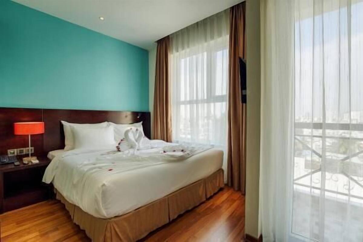 A hotel room with a bed, a nightstand, a red lamp, and a view of the city through large windows.