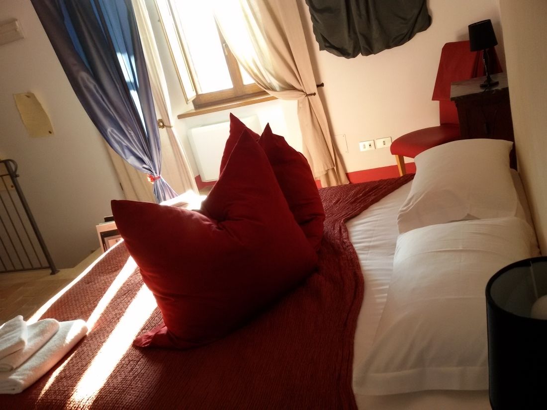 A hotel room with a bed, red pillows, a white pillow, a black lamp, and a window with curtains.