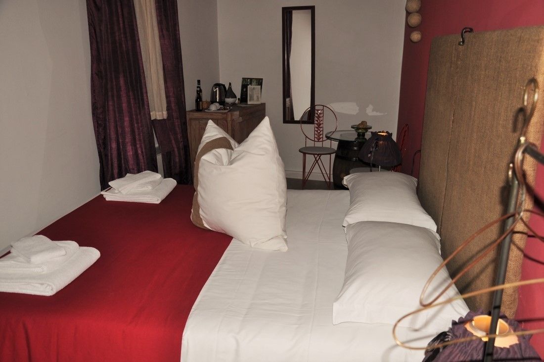 Habitación de hotel con cama, almohadas, colcha roja, cómoda de madera, silla, espejo y cortina.