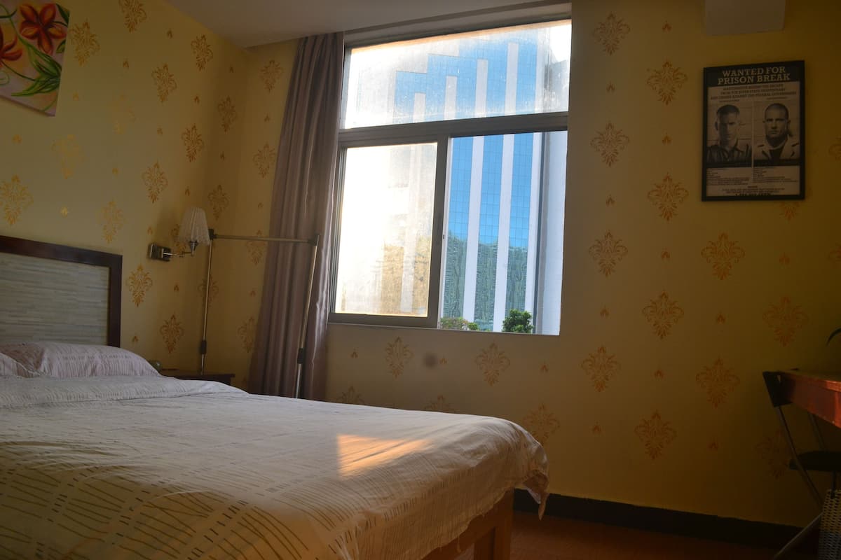A hotel room with a bed, a window with a view of buildings, a poster on the wall, and a lamp.