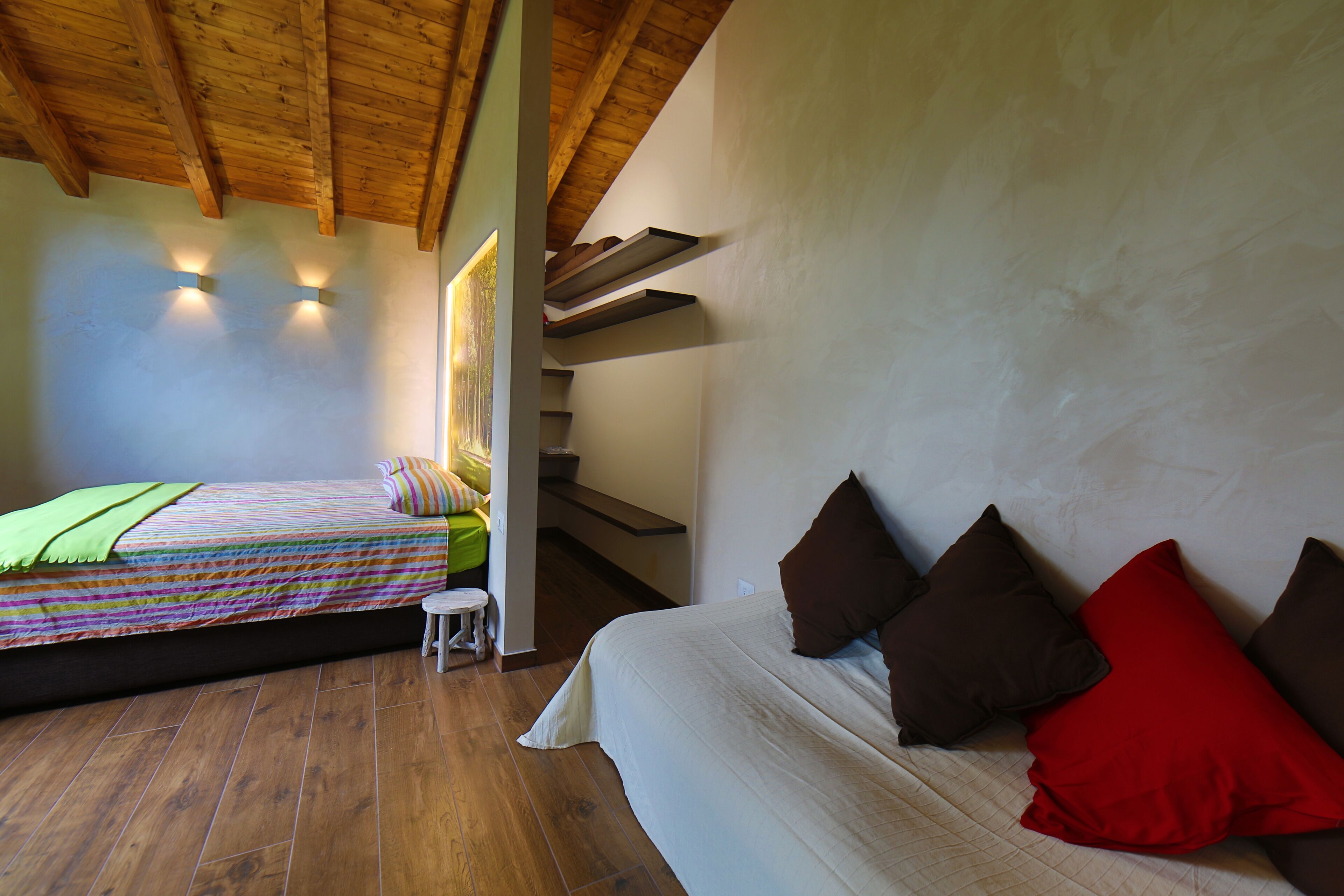 Ein Schlafzimmer mit Holzbalkendecke, einem Bett mit bunter Bettdecke, einem kleinen Hocker und einer Treppe.