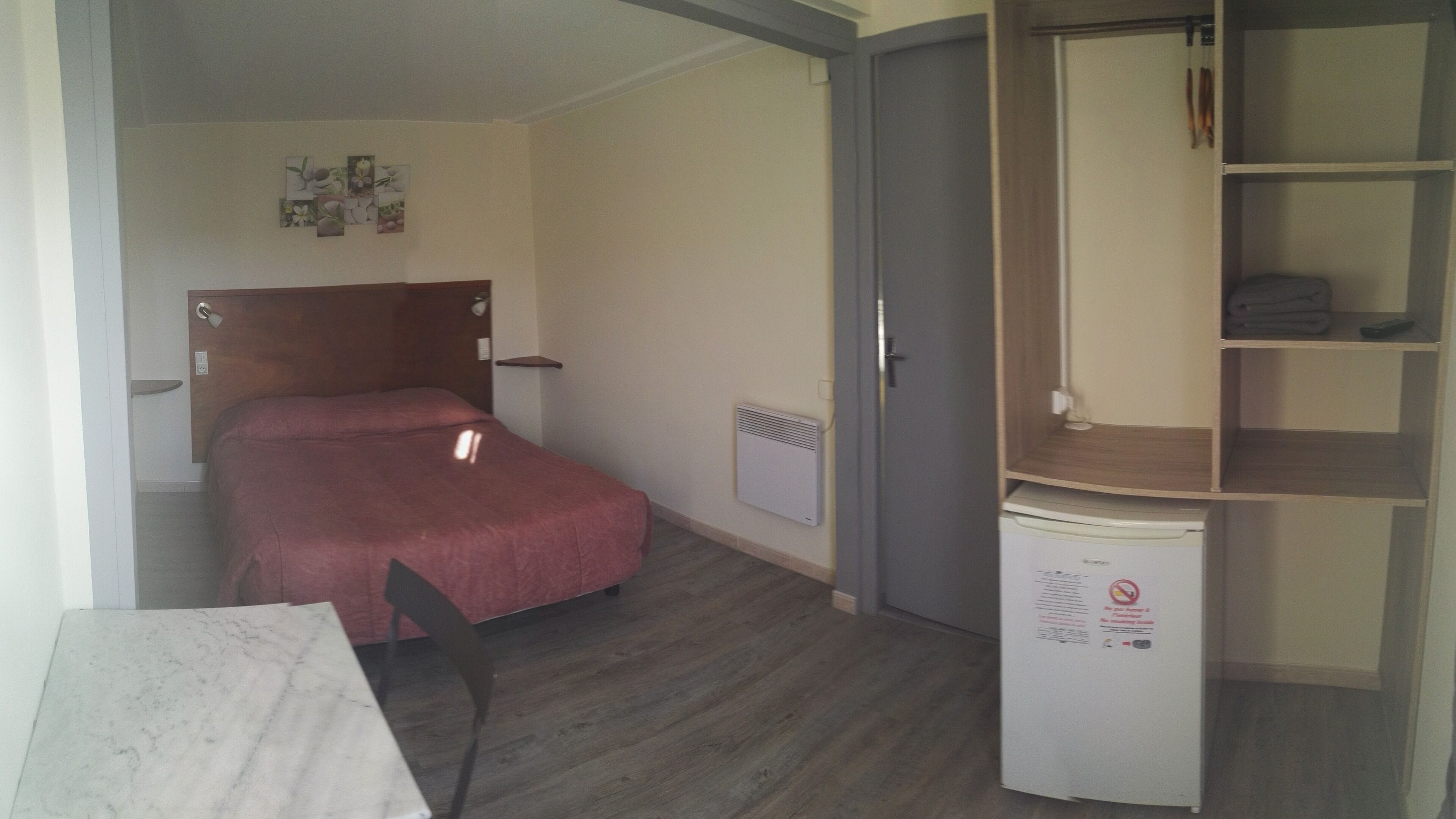 A compact hotel room with a bed, a small table, a chair, a wardrobe, and a built-in heater.