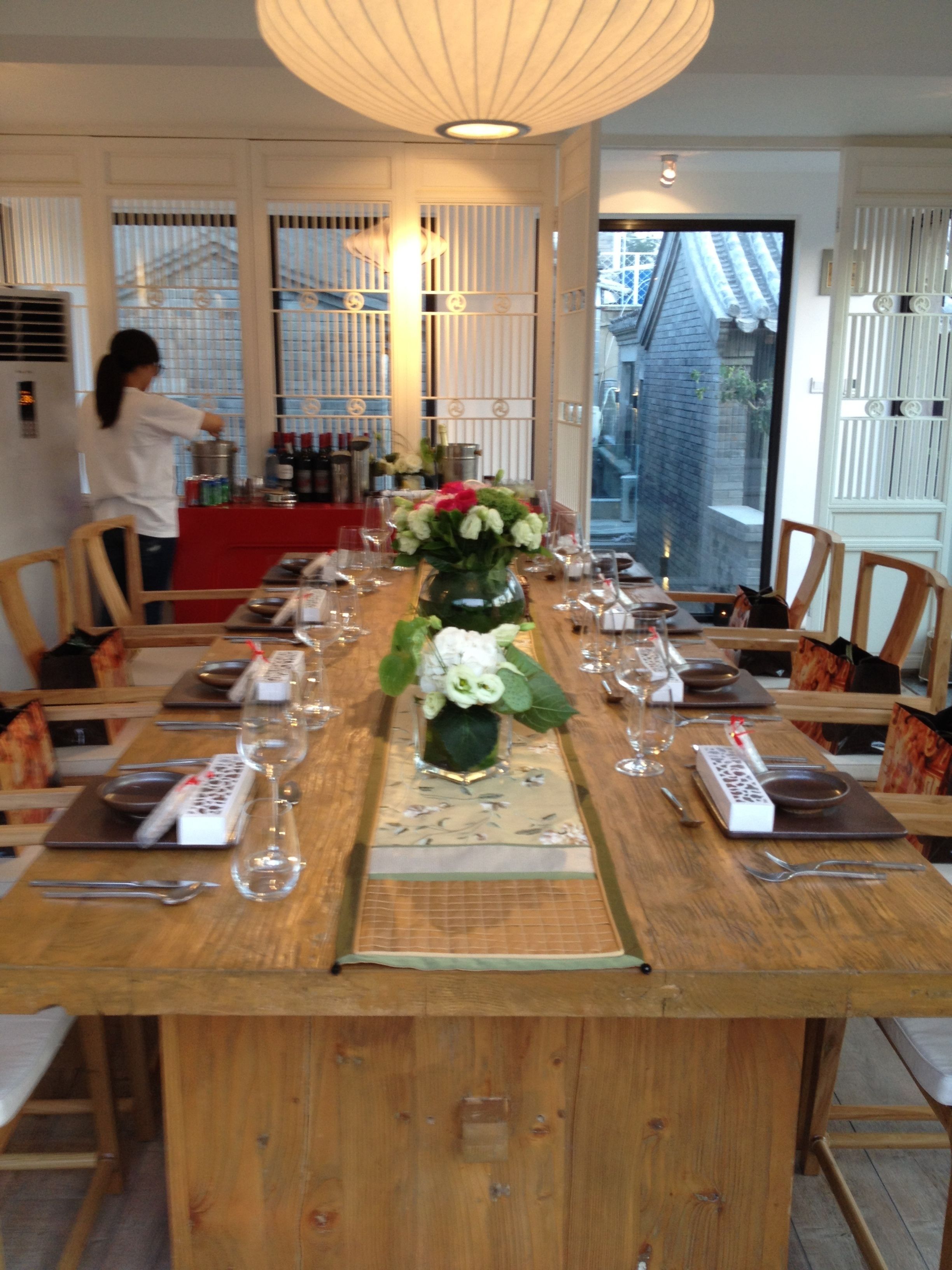 A dining area with a long wooden table set for a meal, a vase with flowers, and a woman in the background.