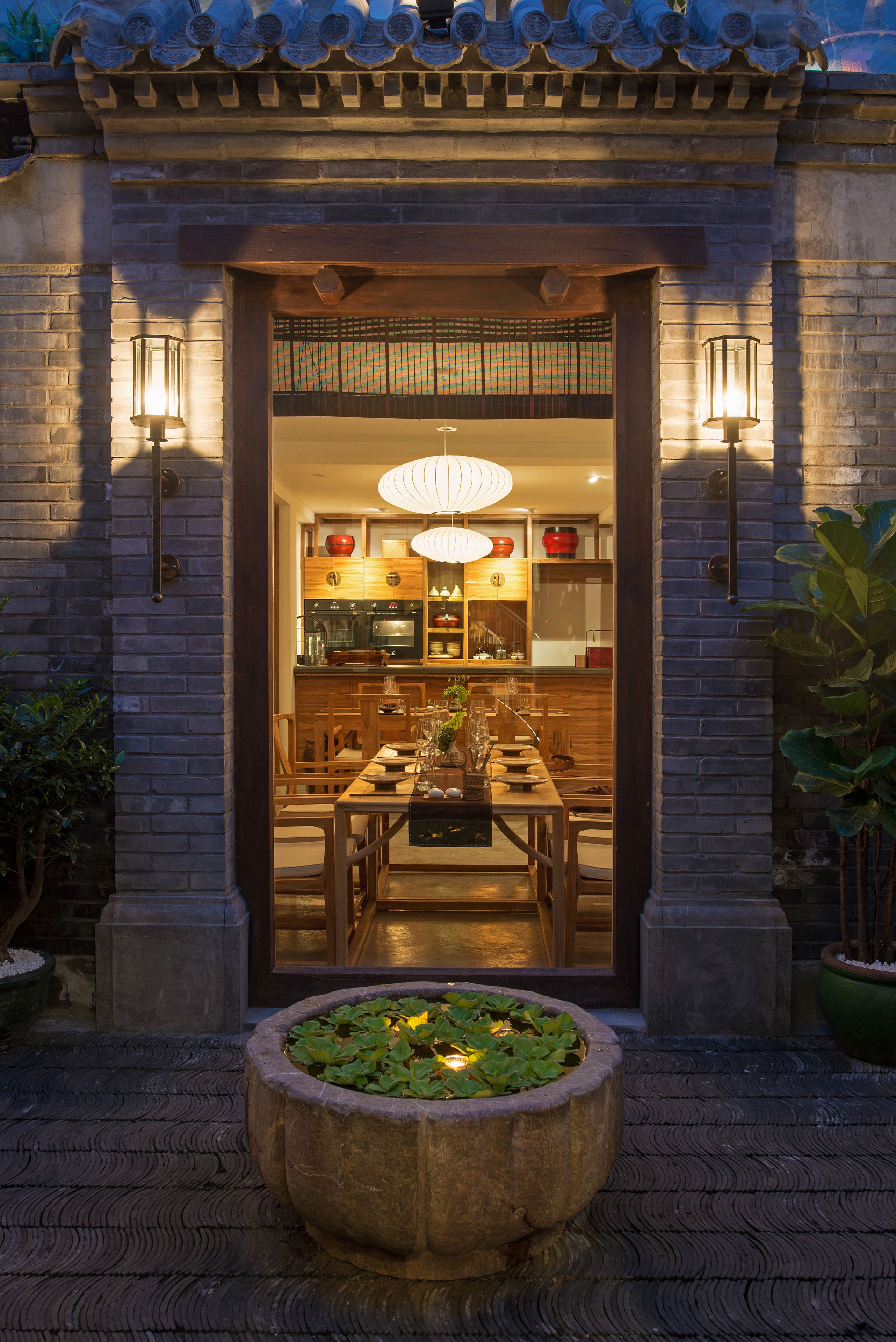 A traditional building entrance with a wooden table and chairs, a hanging light fixture, and a stone planter with plants.