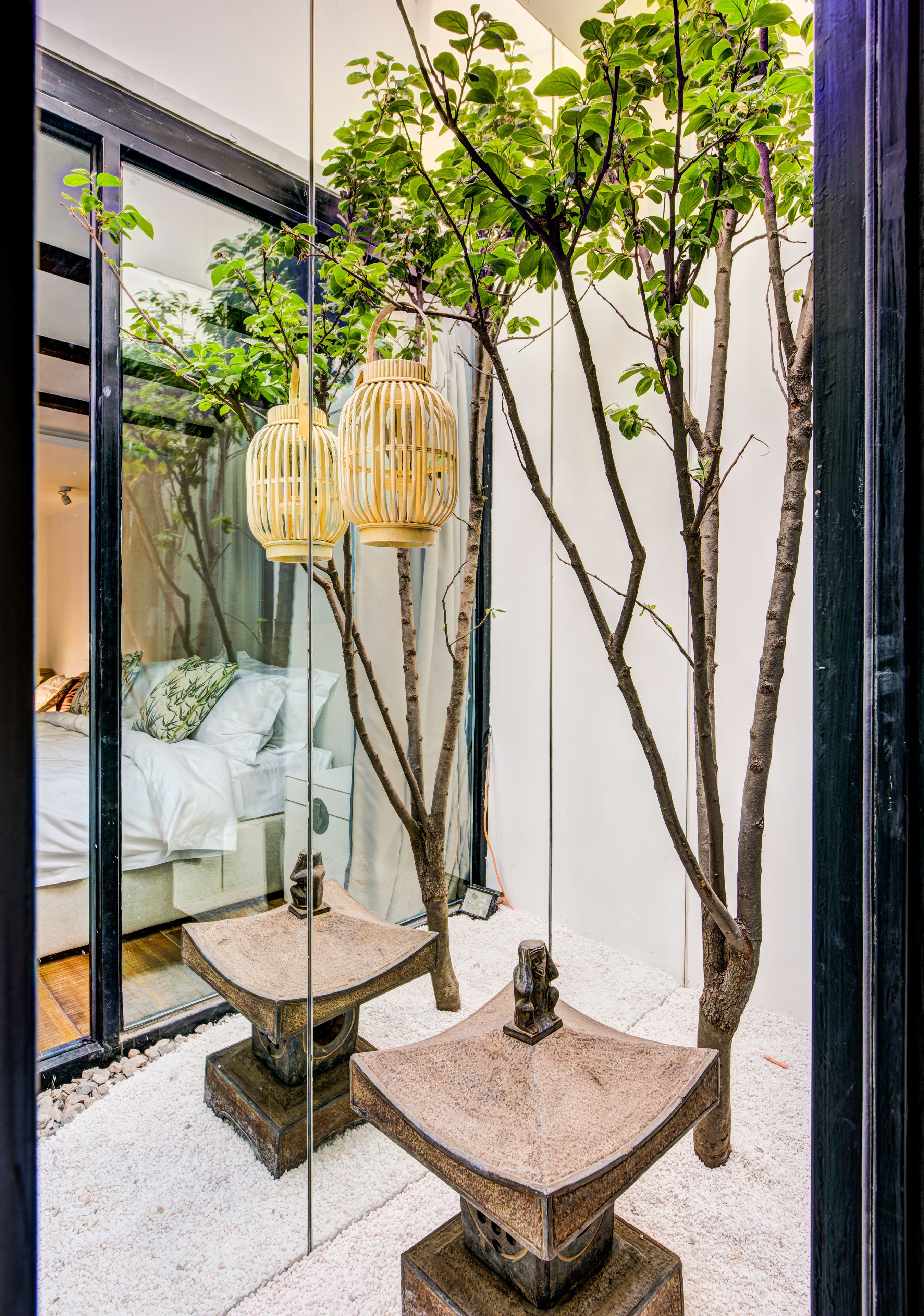 A modern interior with a glass door reflecting a bedroom, a small tree, and two stone tables with hanging lanterns.