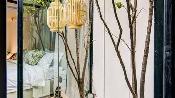 A modern interior with a glass door reflecting a bedroom, a small tree, and two stone tables with hanging lanterns.