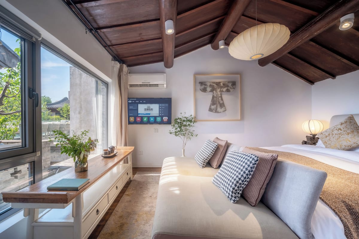 A modern bedroom with a large window, a bedside table with a plant, a mounted TV, and a bed with multiple pillows.