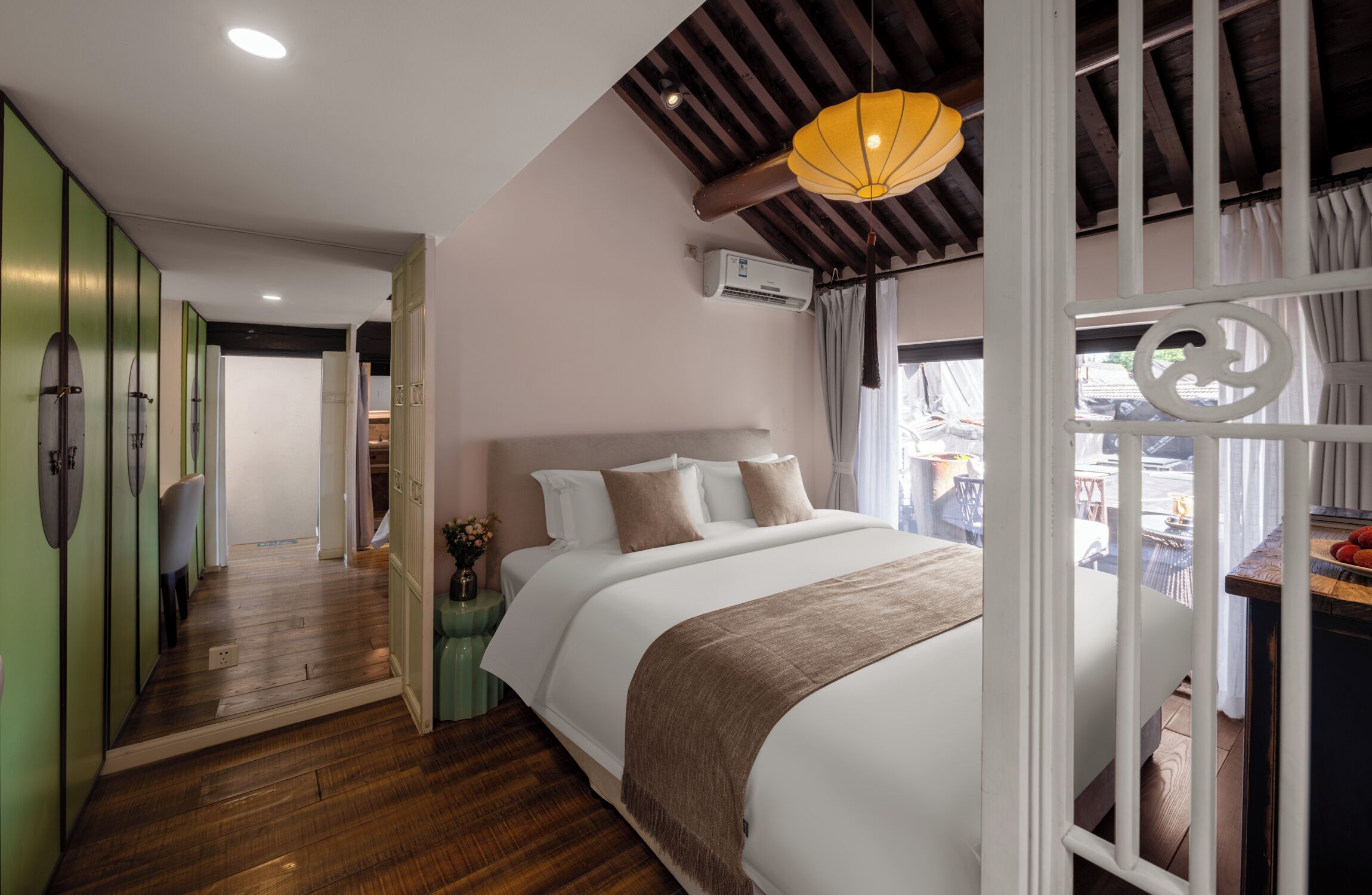 A bedroom with a large bed, wooden flooring, a ceiling fan, and a view of the outside.