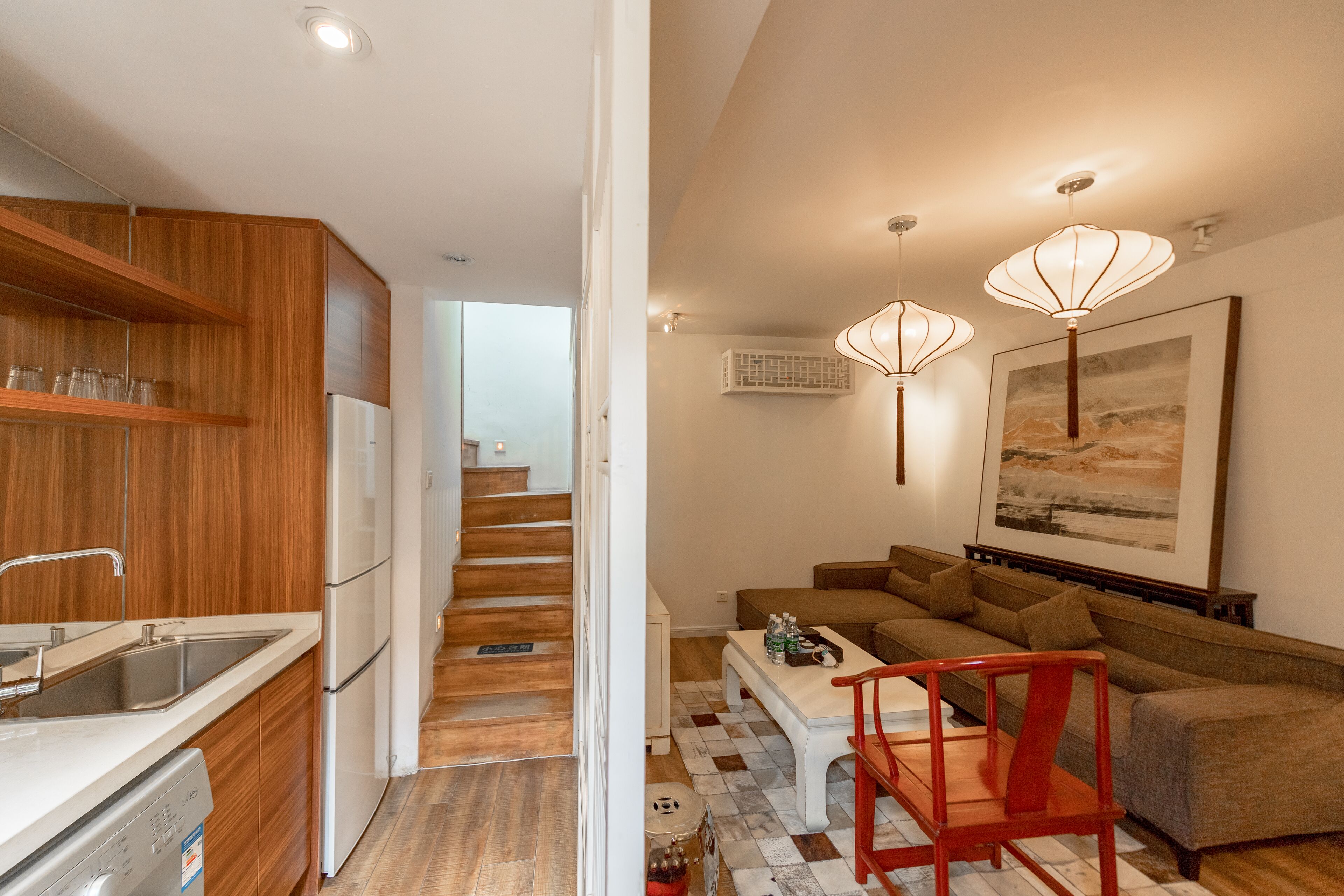 A modern kitchen with wooden cabinets, a white refrigerator, and a built-in dishwasher. There is a staircase leading to another level. The living area features a brown sofa, a white coffee table, and a red chair. Pendant lights hang above the sofa.
