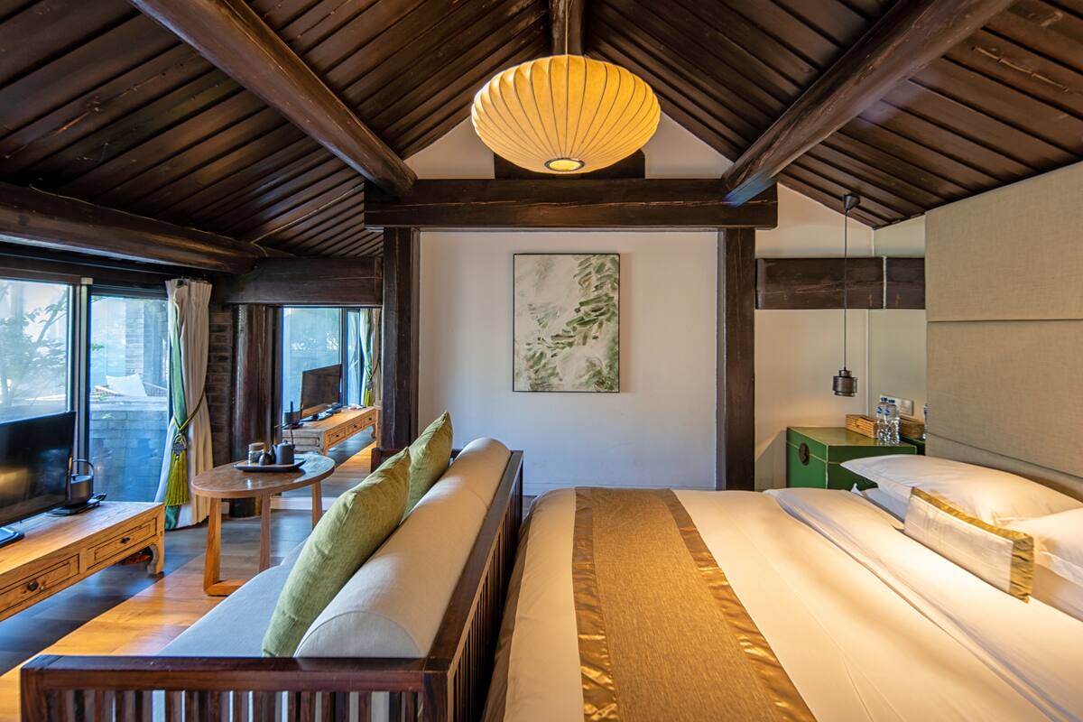 A modern hotel room with a wooden ceiling, a large bed, a sofa, a small table, and a painting on the wall.