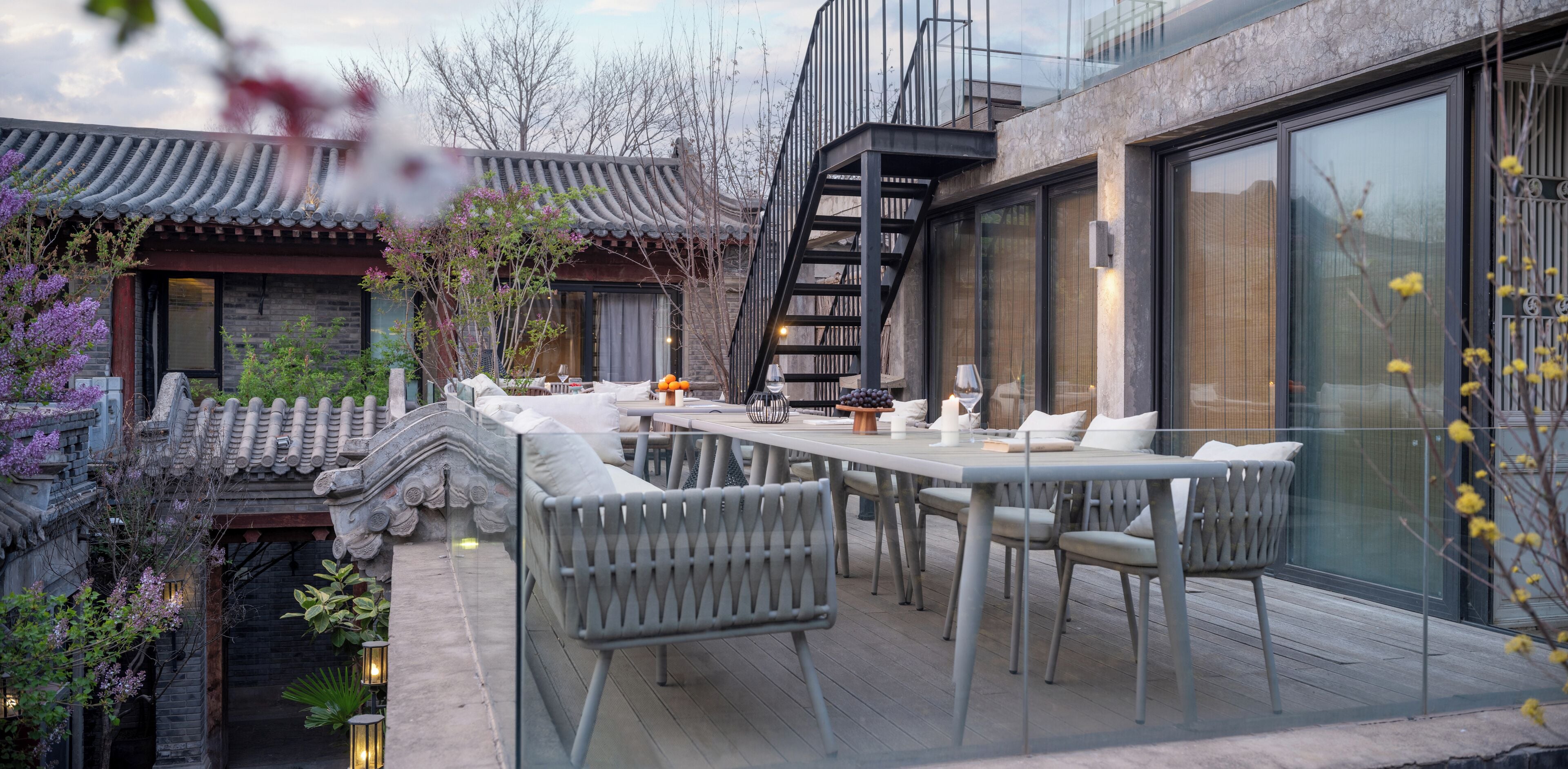 Un área de comedor al aire libre moderna con terraza de madera, mobiliario blanco y un edificio de estilo tradicional al fondo.