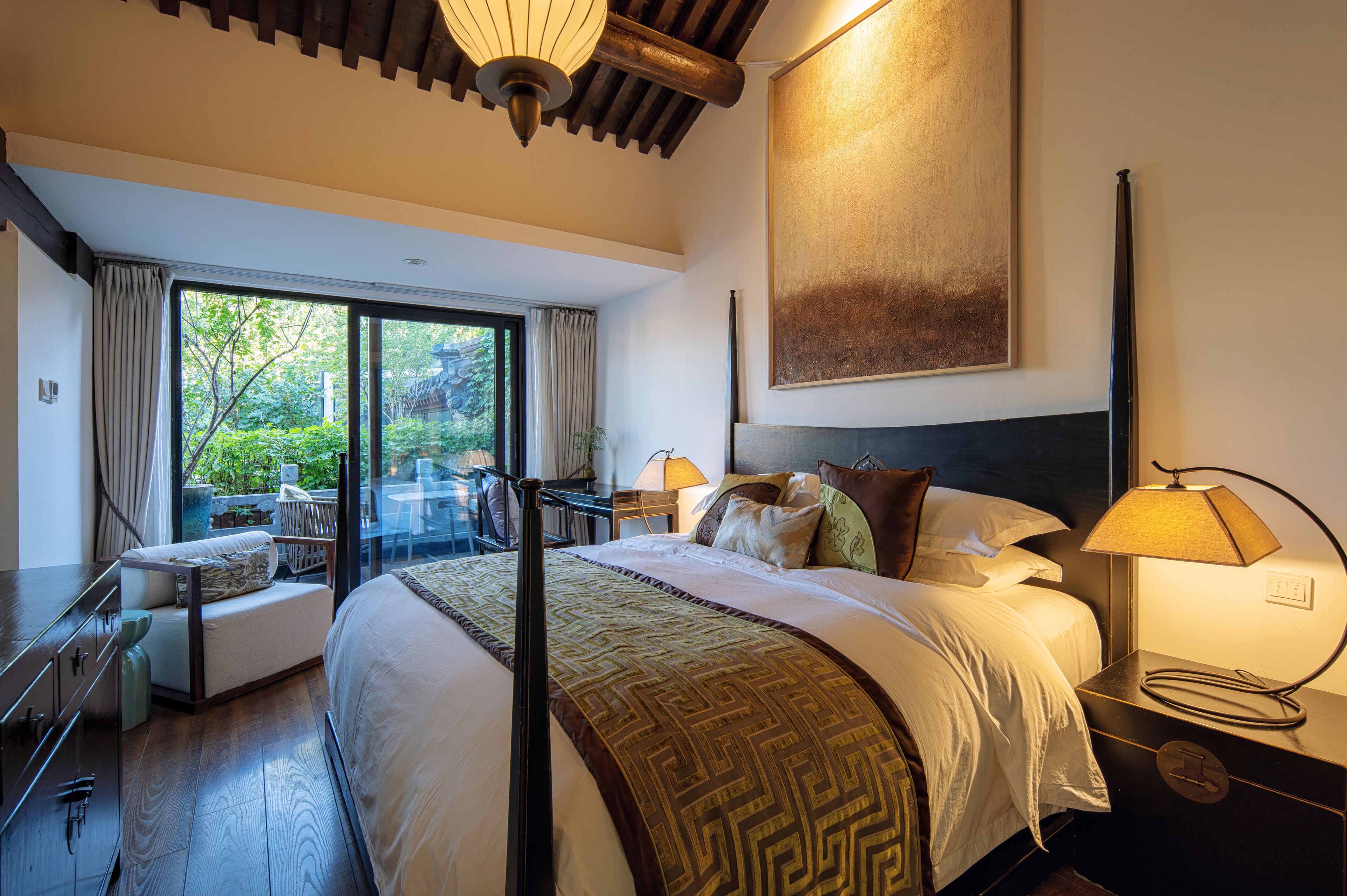 A spacious bedroom with a large bed, a sitting area with a chair and table, and a view of greenery outside.