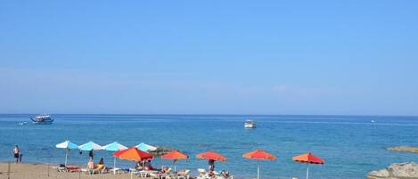 A beach with rows of sun loungers and umbrellas, a clear blue sky, and boats on the water.